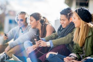 A multi-ethnic group of college age students are sitting together texting on their cell phones and taking selfies together.